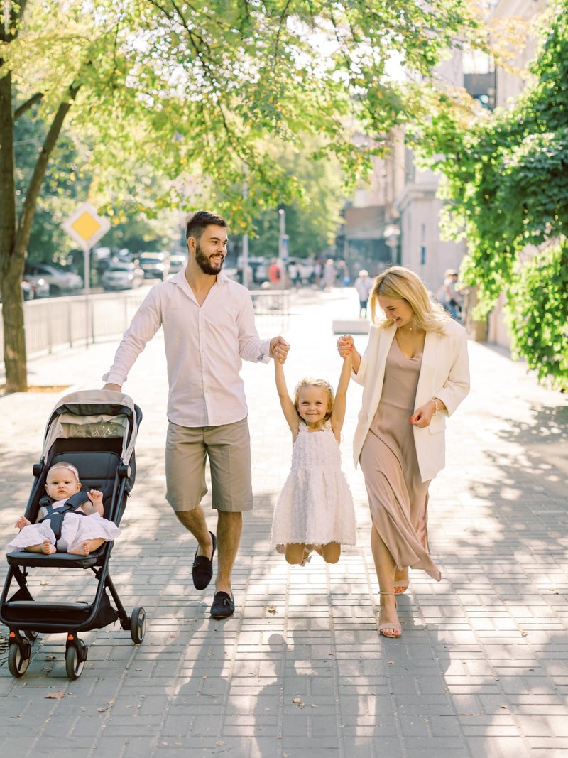 Family walking together with a stroller on a tree-lined sidewalk.